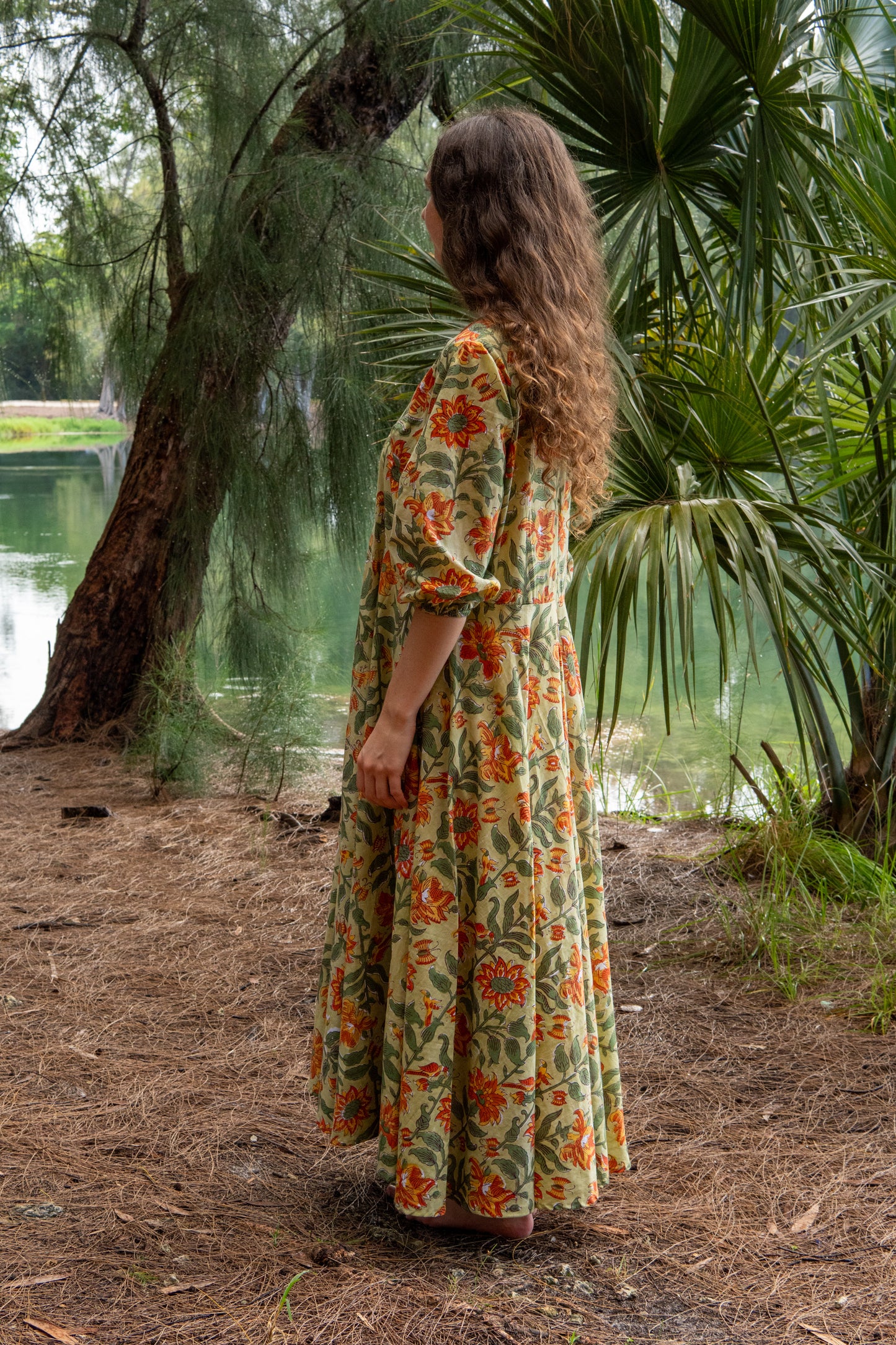 Woman in a floral dress standing in a natural setting with trees and water.