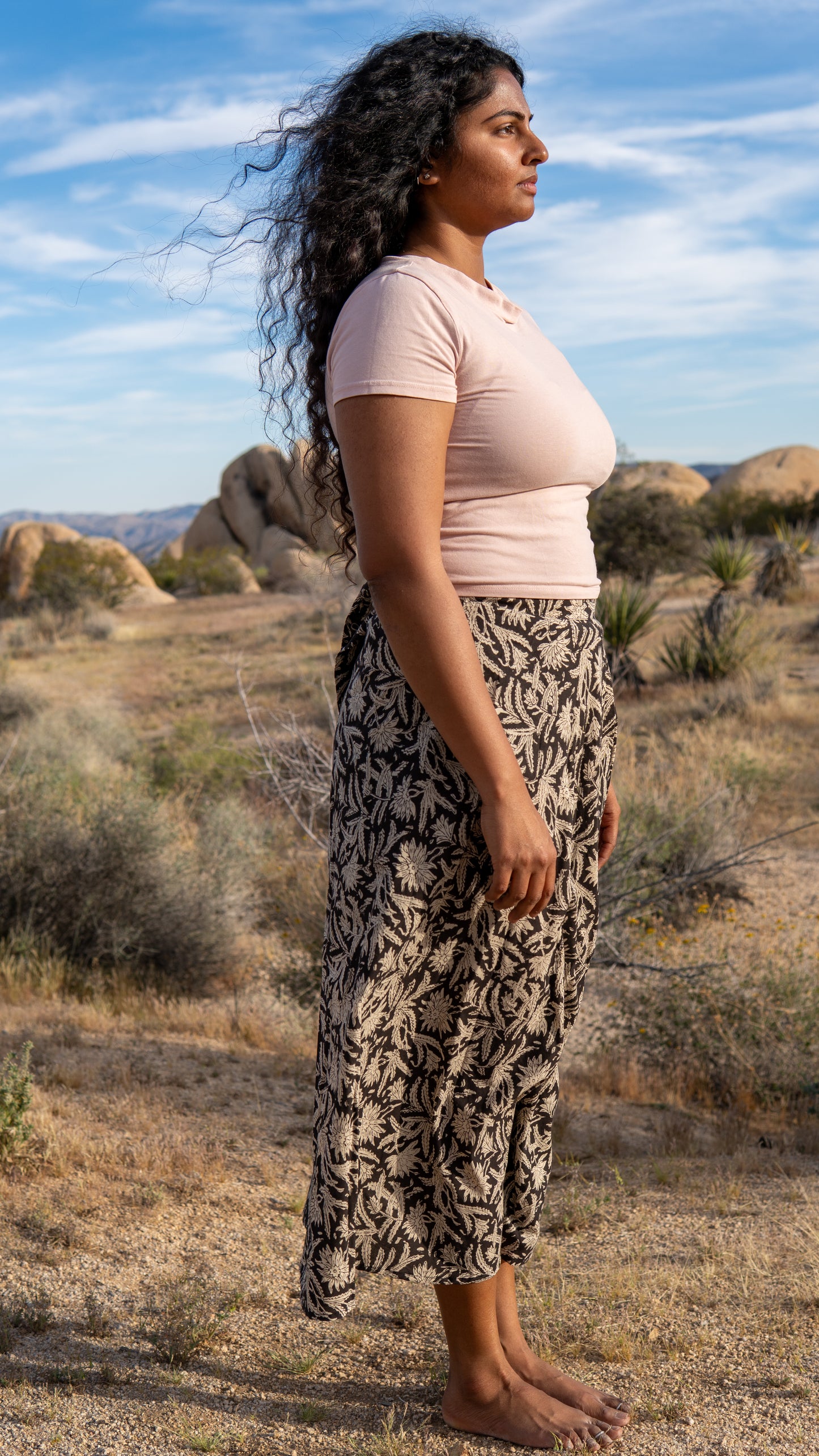 Woman standing in a desert landscape wearing a pink top and patterned pants.