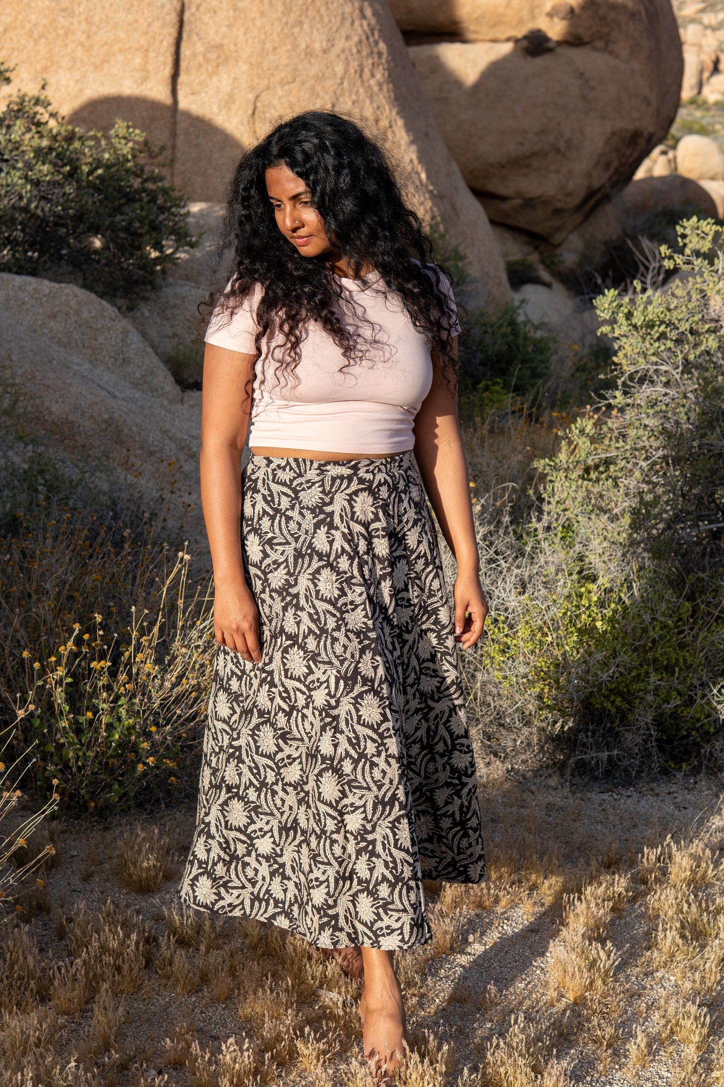 Woman standing in a desert landscape with large rocks and sparse vegetation.