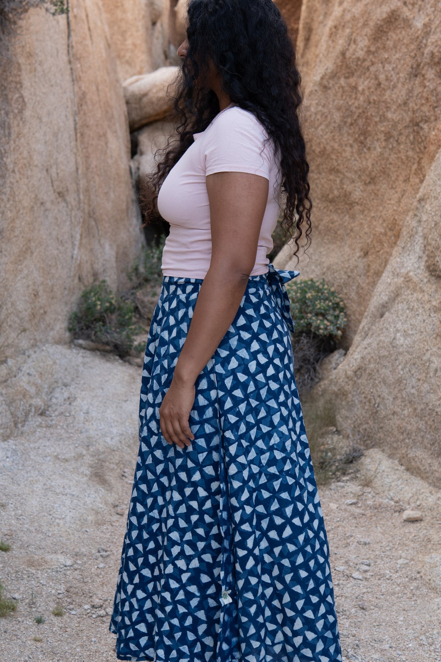 Woman in a white top and blue patterned skirt standing in a rocky desert landscape.