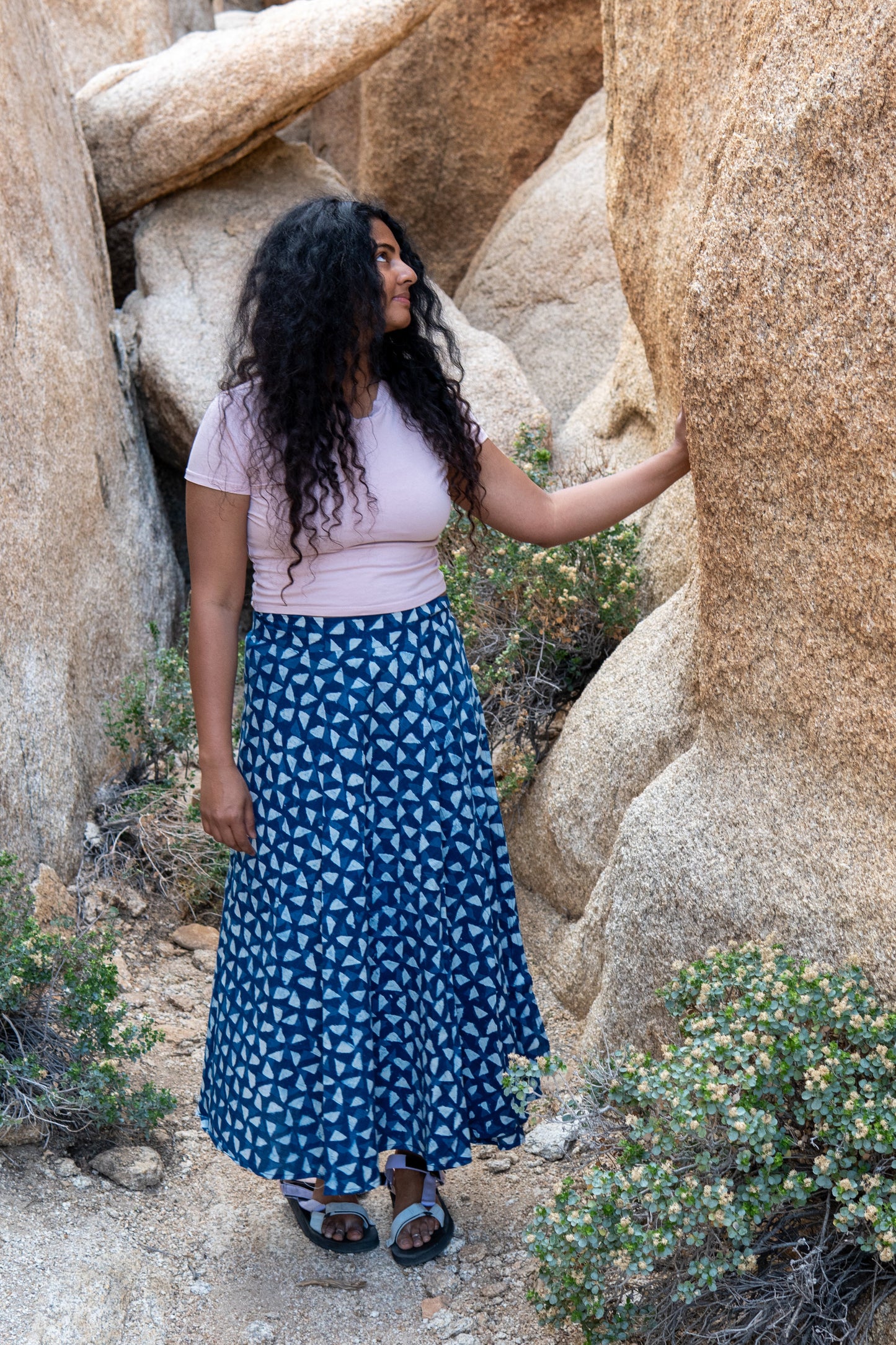 Woman in a pink top and blue skirt standing among large rocks in a desert setting