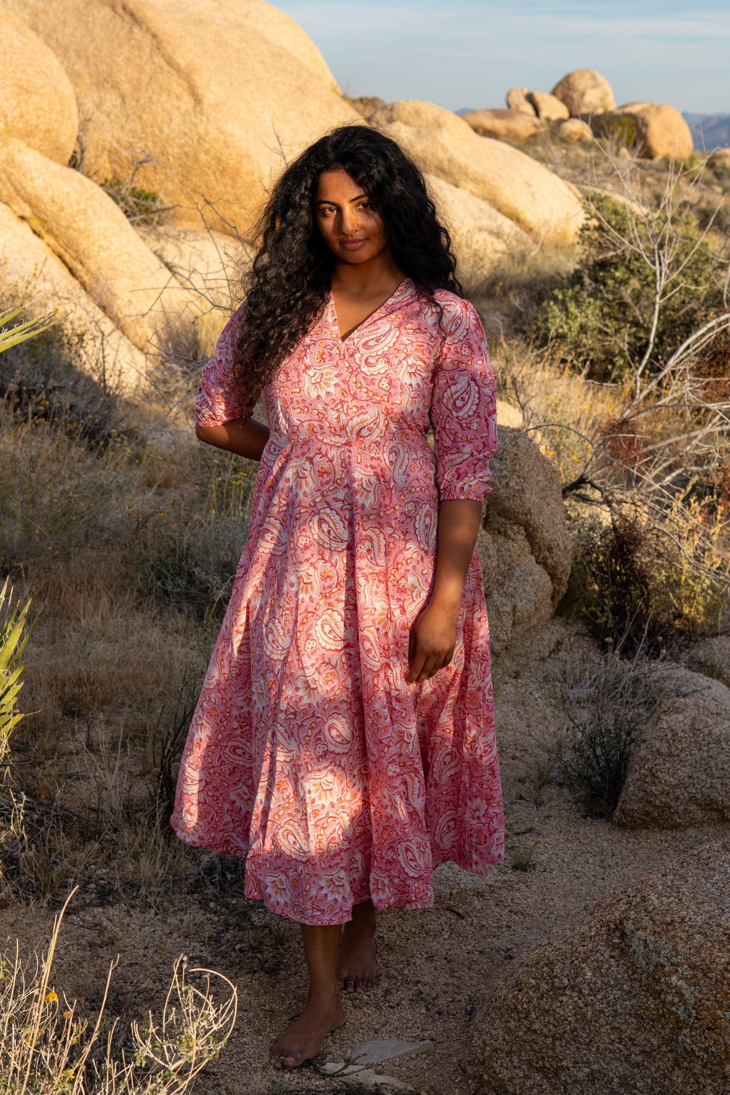 Woman in a pink dress standing in a desert landscape with large rocks and sparse vegetation.