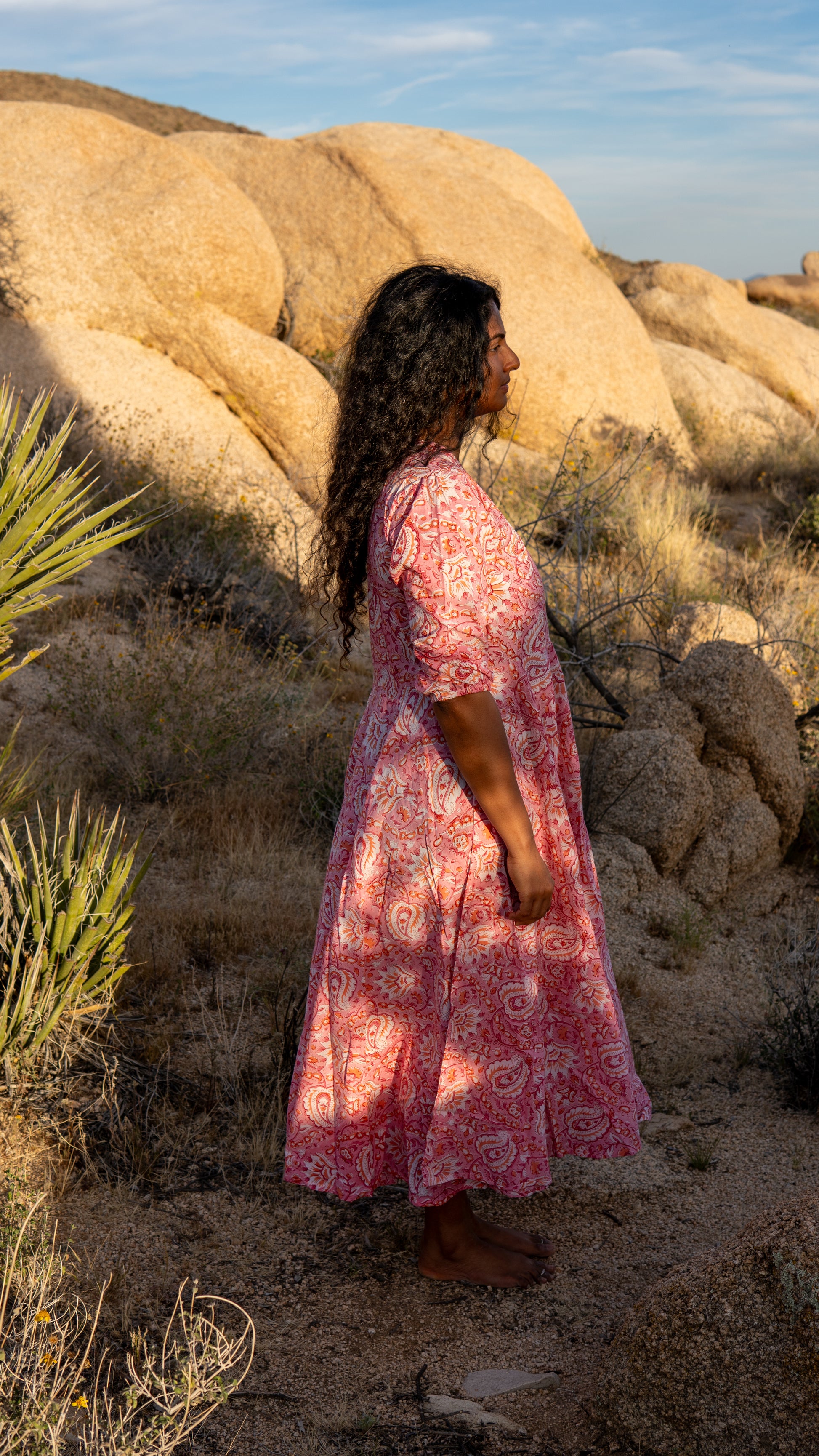 Woman in a pink dress standing in a desert landscape with large rocks and sparse vegetation.