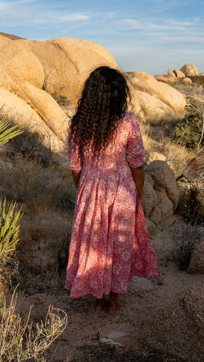 Woman in a pink dress standing in a desert landscape with large rocks and sparse vegetation.