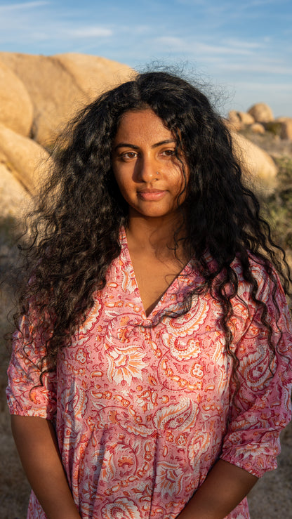 Woman in a pink patterned shirt standing in a desert landscape with large rocks and blue sky.