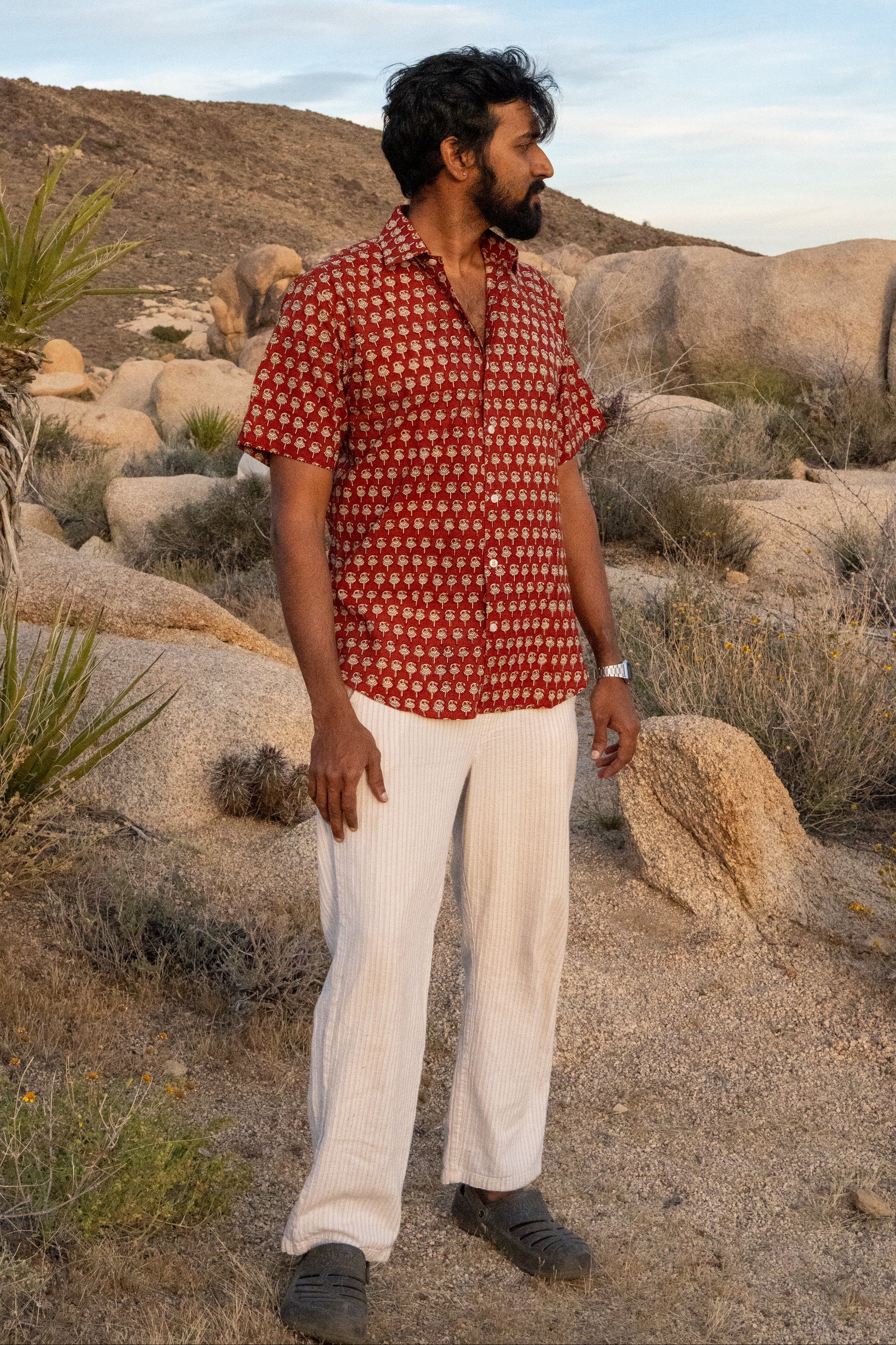 Man in a red patterned shirt and white pants standing in a desert landscape with rocks and sparse vegetation.