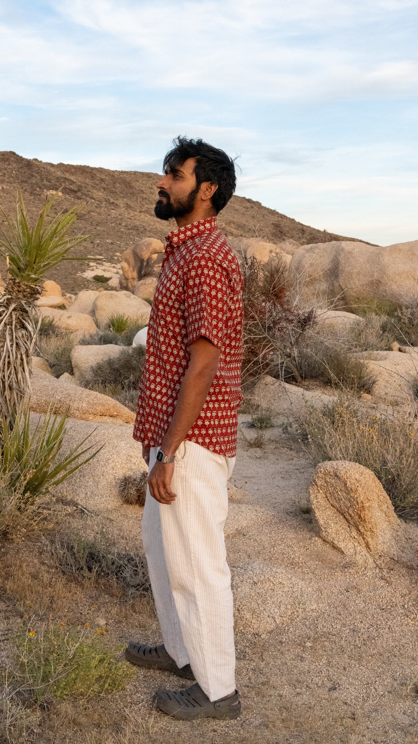 Man standing in a desert landscape wearing a red patterned shirt and white pants.