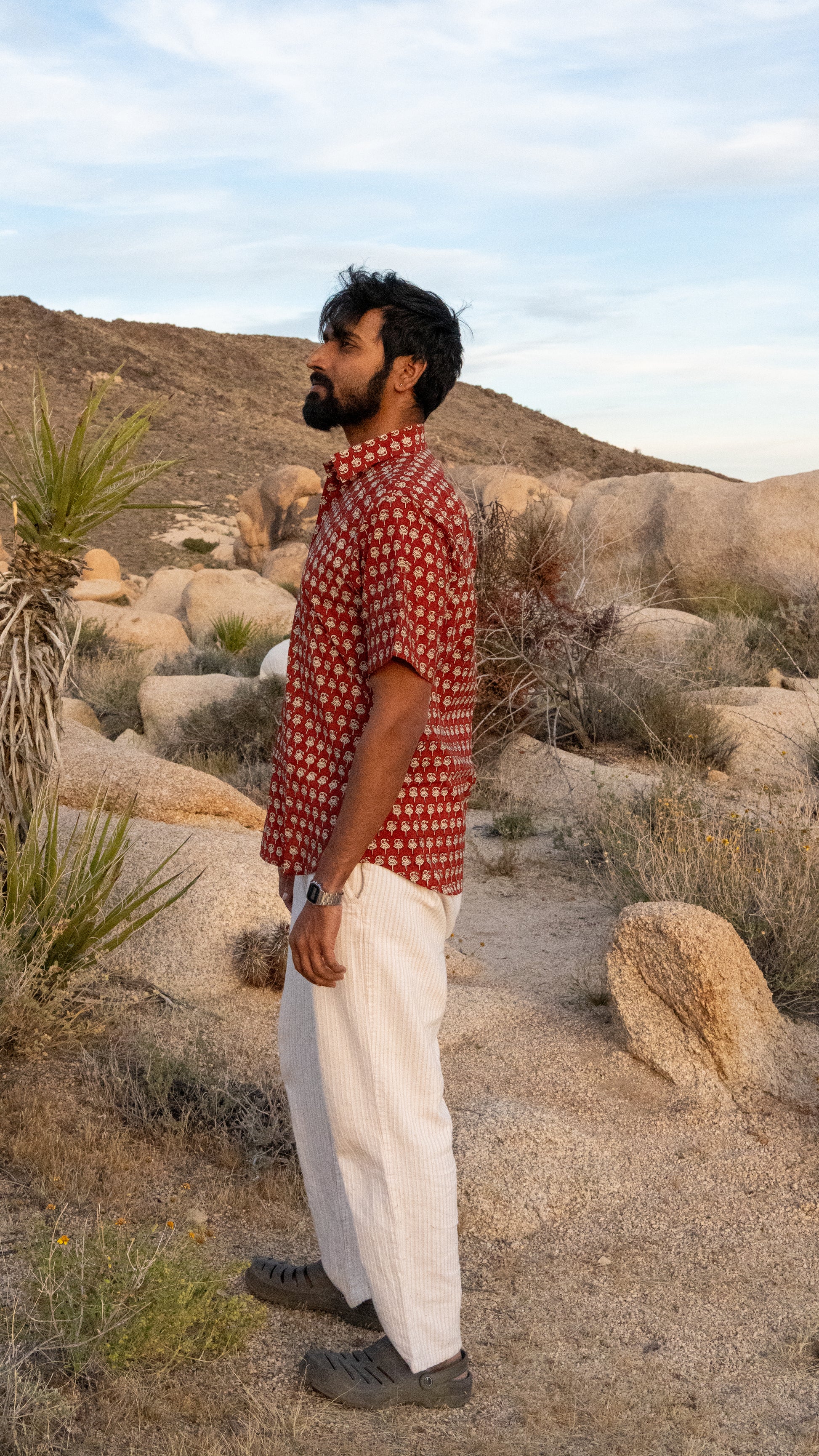 Man standing in a desert landscape wearing a red patterned shirt and white pants.