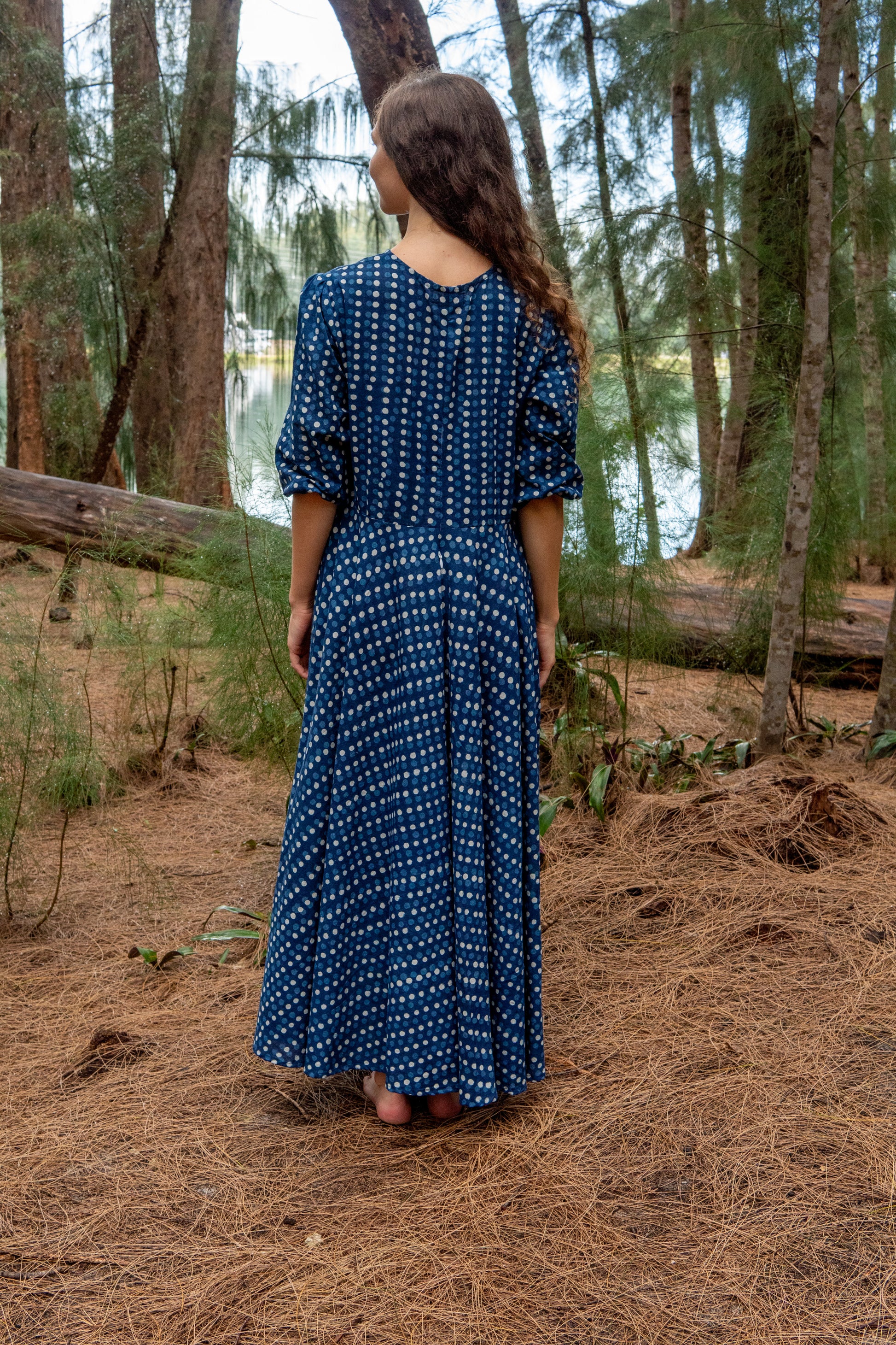Woman in an indigo blue dress standing in a forest with trees and water in the background