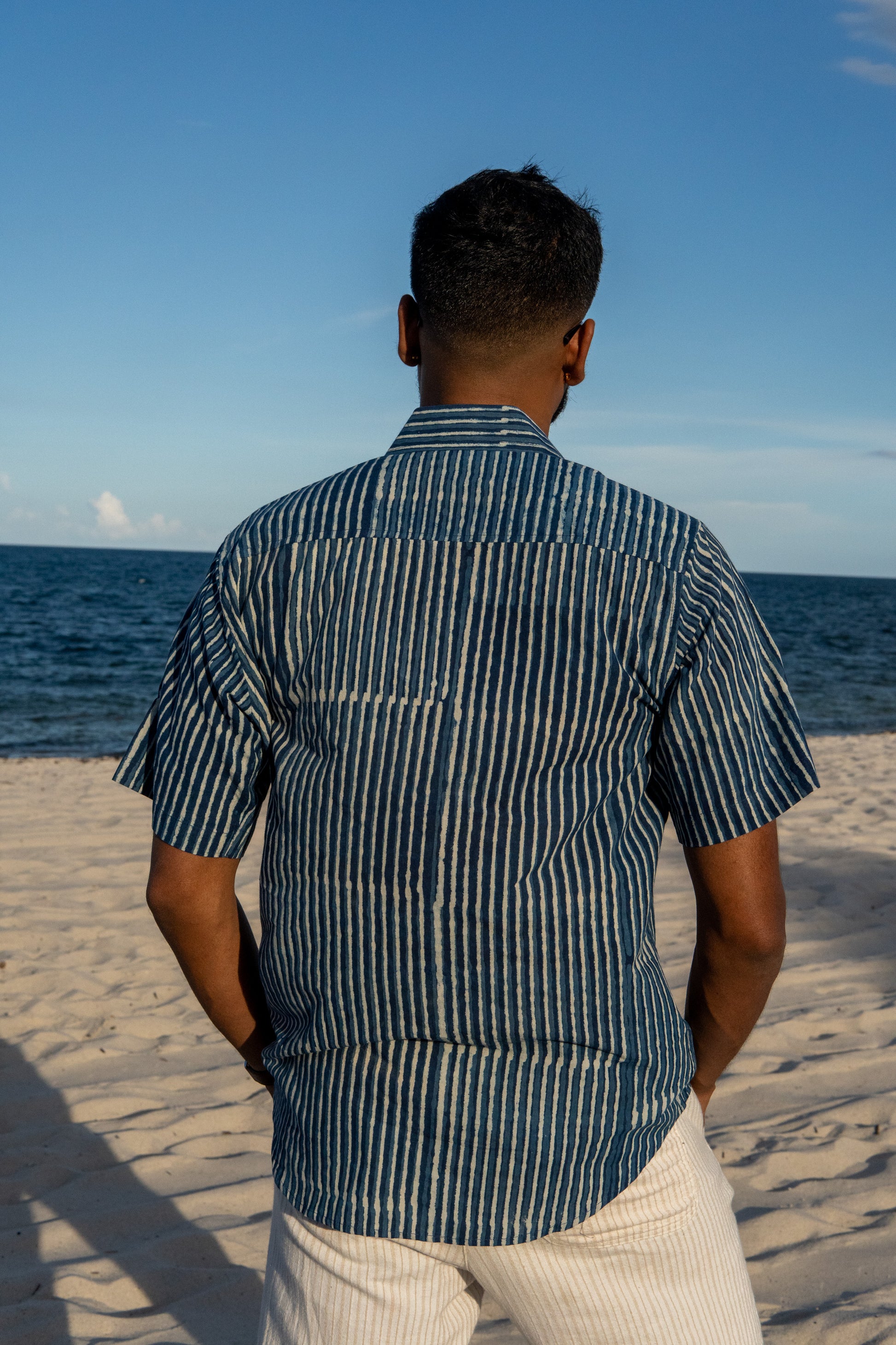 Man standing on a beach wearing a striped shirt, looking at the ocean.
