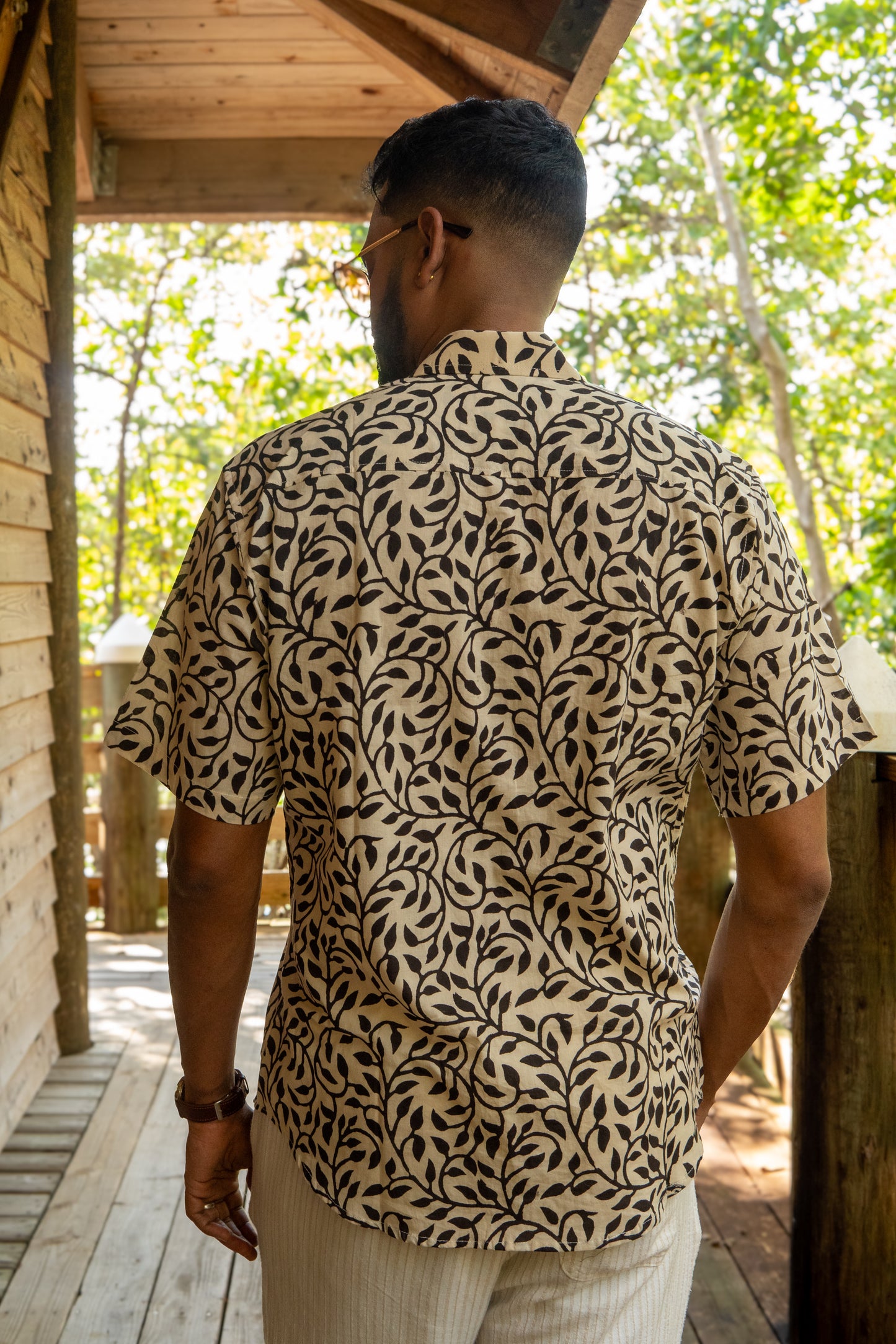 Man wearing a patterned shirt standing on a wooden deck with greenery in the background