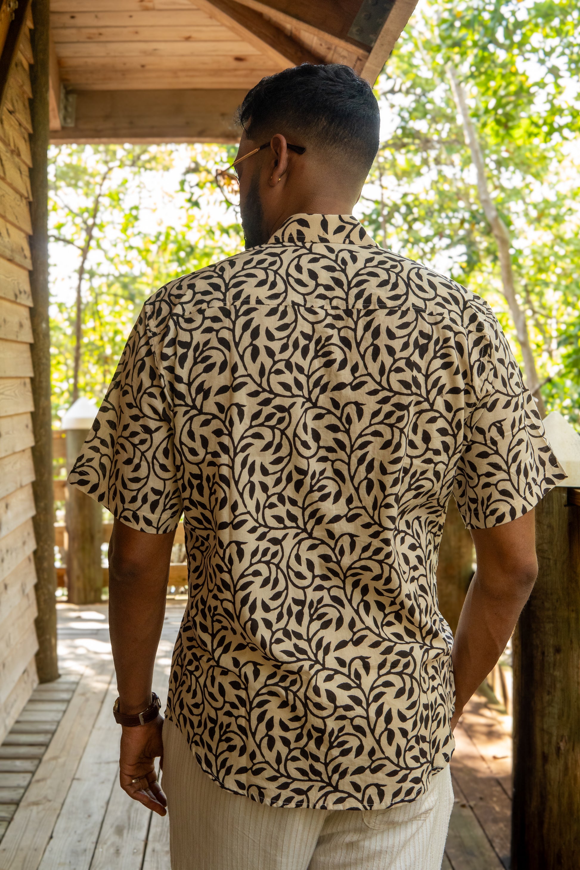 Man wearing a patterned shirt standing on a wooden deck with greenery in the background