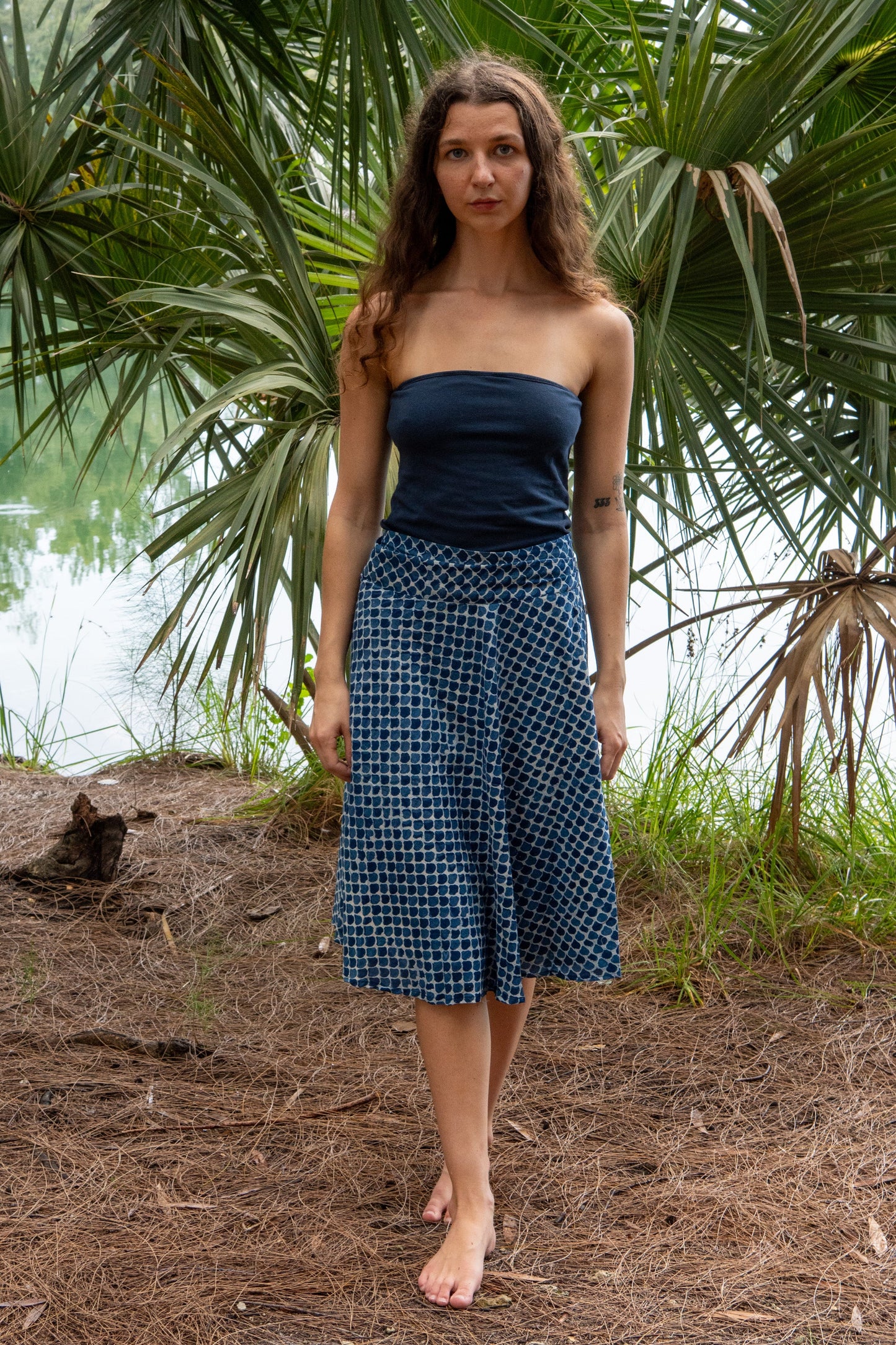 Woman wearing a blue strapless top and checkered skirt standing in a natural setting with greenery.