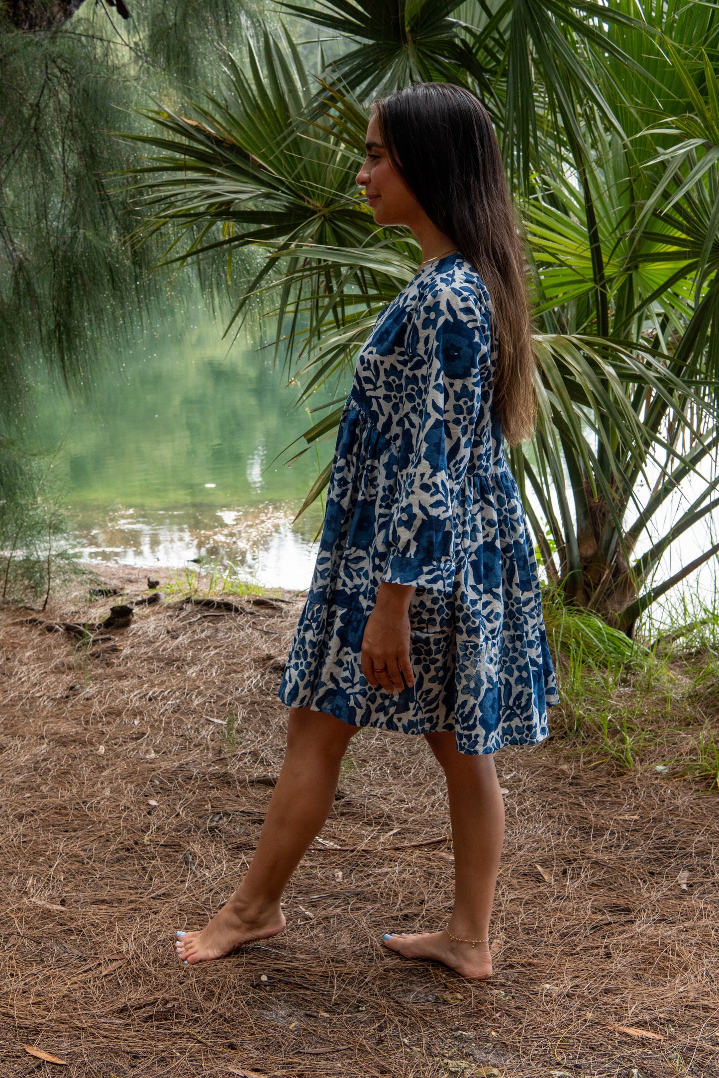 Woman in a blue floral dress walking in a natural setting with greenery and water.