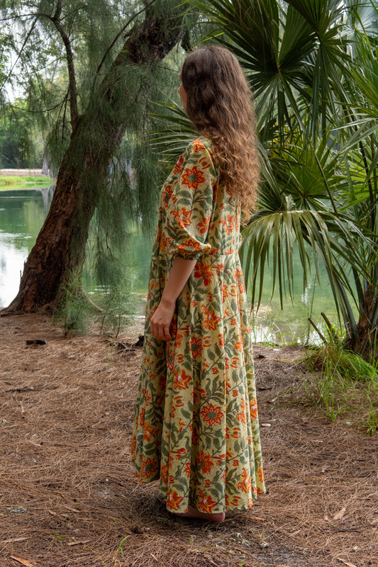 Woman in a floral dress standing in a natural setting with trees and water.